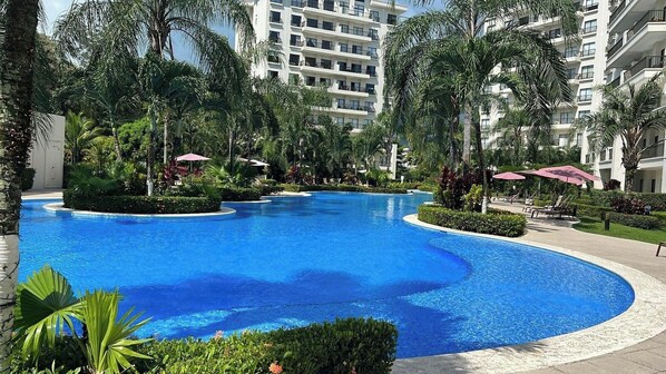 A large, curved swimming pool surrounded by lush greenery and palm trees, with a modern building in the background.