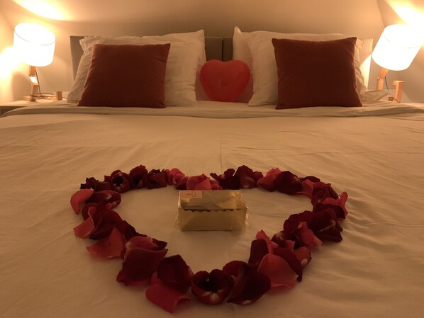 Una cama con sábanas blancas, un arreglo de pétalos de rosa en forma de corazón y una pequeña caja en el centro.