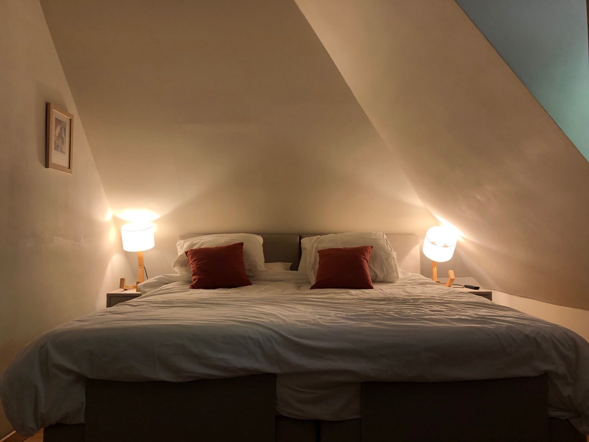 Een slaapkamer met een schuine kap, twee bedlampen, een bed met witte lakens en twee rode kussens, en een lijst met een afbeelding aan de muur.