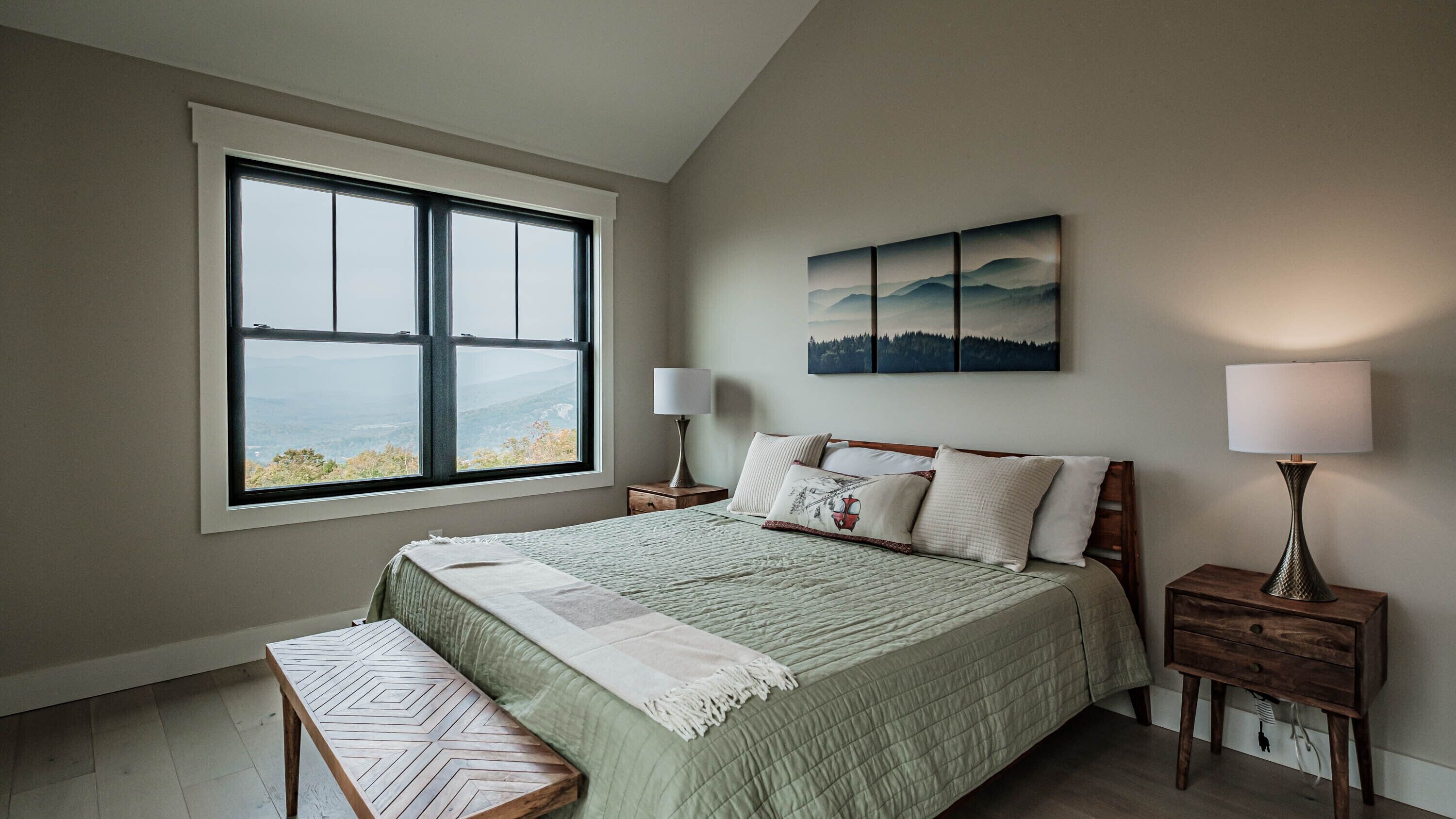 A bedroom with a large window, a bed with multiple pillows, two bedside tables with lamps, and a wall-mounted painting.