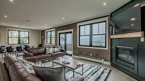 A spacious living room with a large sofa, a coffee table, and a fireplace. There are large windows offering a view of the outdoors. - Panoramic mountain view/Hot Tub/Shuffleboard/Theater room  Sunday River (Newry)