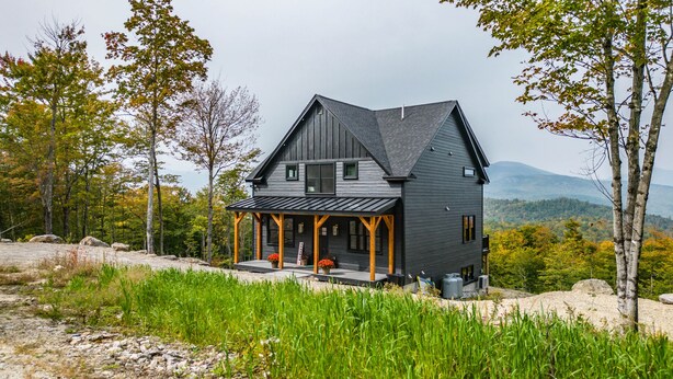 A two-story house with a dark gray exterior, a covered porch, and a view of a mountainous landscape. - Panoramic mountain view/Hot Tub/Shuffleboard/Theater room  Sunday River (Newry)