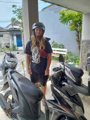 A man standing next to two motorcycles in an outdoor area with a building and some greenery in the background. - CALDERA PARK HOMESTAY SYARIAH (Probolinggo)
