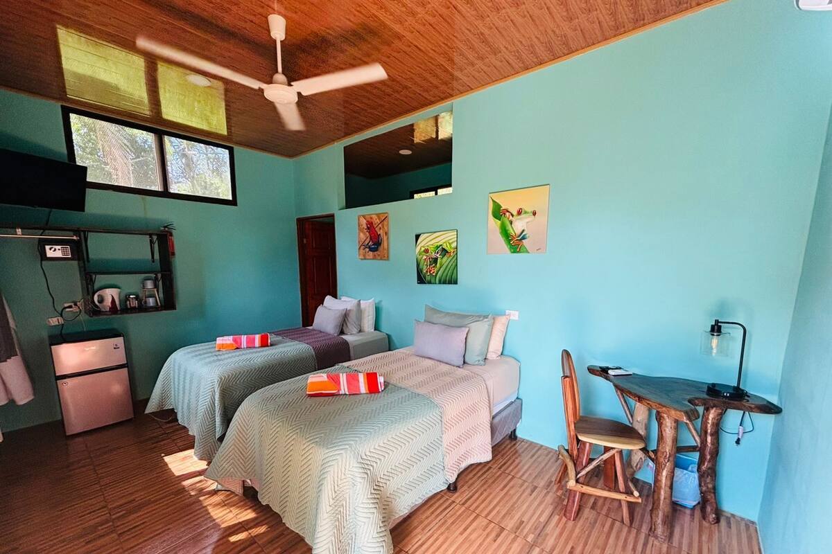 A room with two beds, a desk, a chair, a ceiling fan, and a refrigerator.