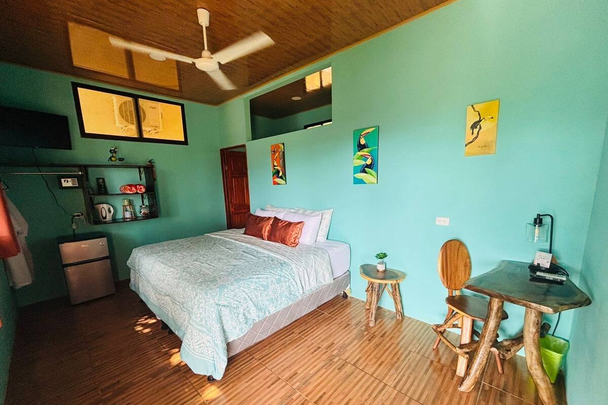 A bedroom with a bed, a desk, a chair, a small table, a ceiling fan, and a shelf with various items.