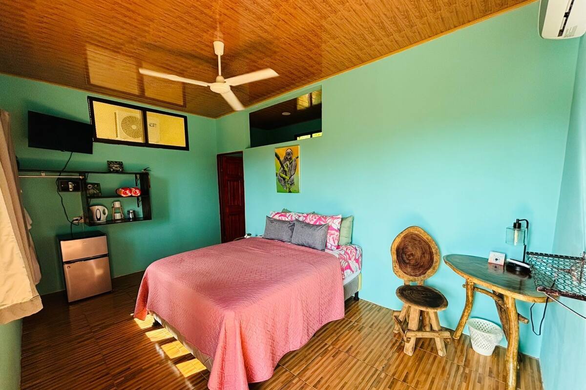 A bedroom with a bed, a desk, a chair, a ceiling fan, a TV, and a refrigerator.