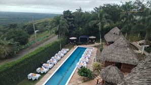 Un área de piscina con sillones de descanso y sombrillas, rodeada de abundante vegetación y estructuras con techo de paja.