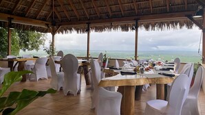 Un área de comedor al aire libre con mesas y sillas, un techo de paja y vistas del paisaje.