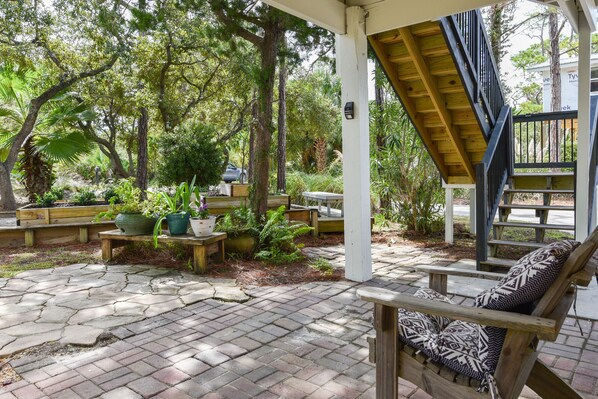 Un patio techado con un banco y una silla de madera, rodeado de plantas en macetas y árboles.