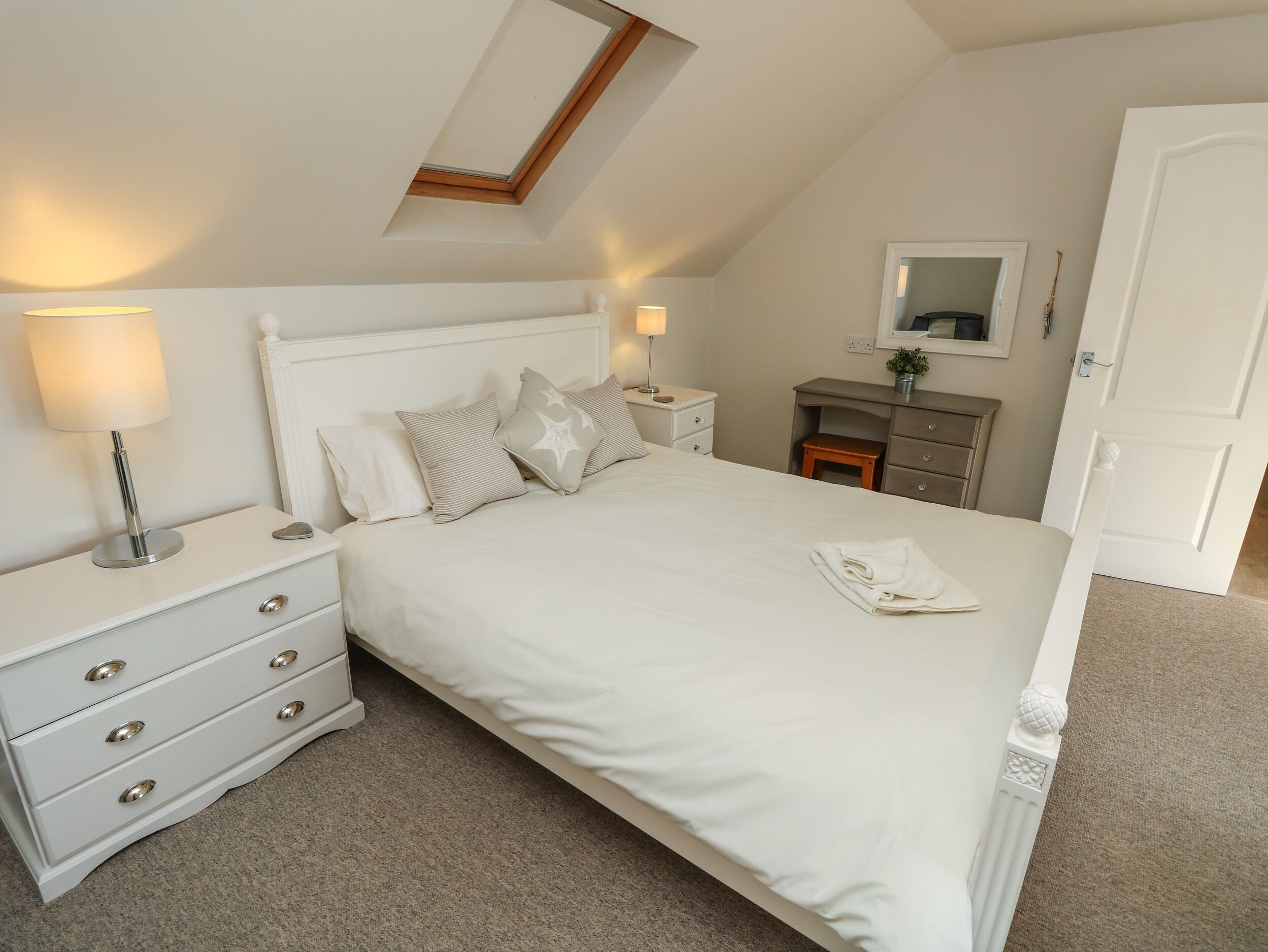 A neatly arranged bedroom with a bed, bedside tables, a nightstand, a dresser, a mirror, and a small stool.