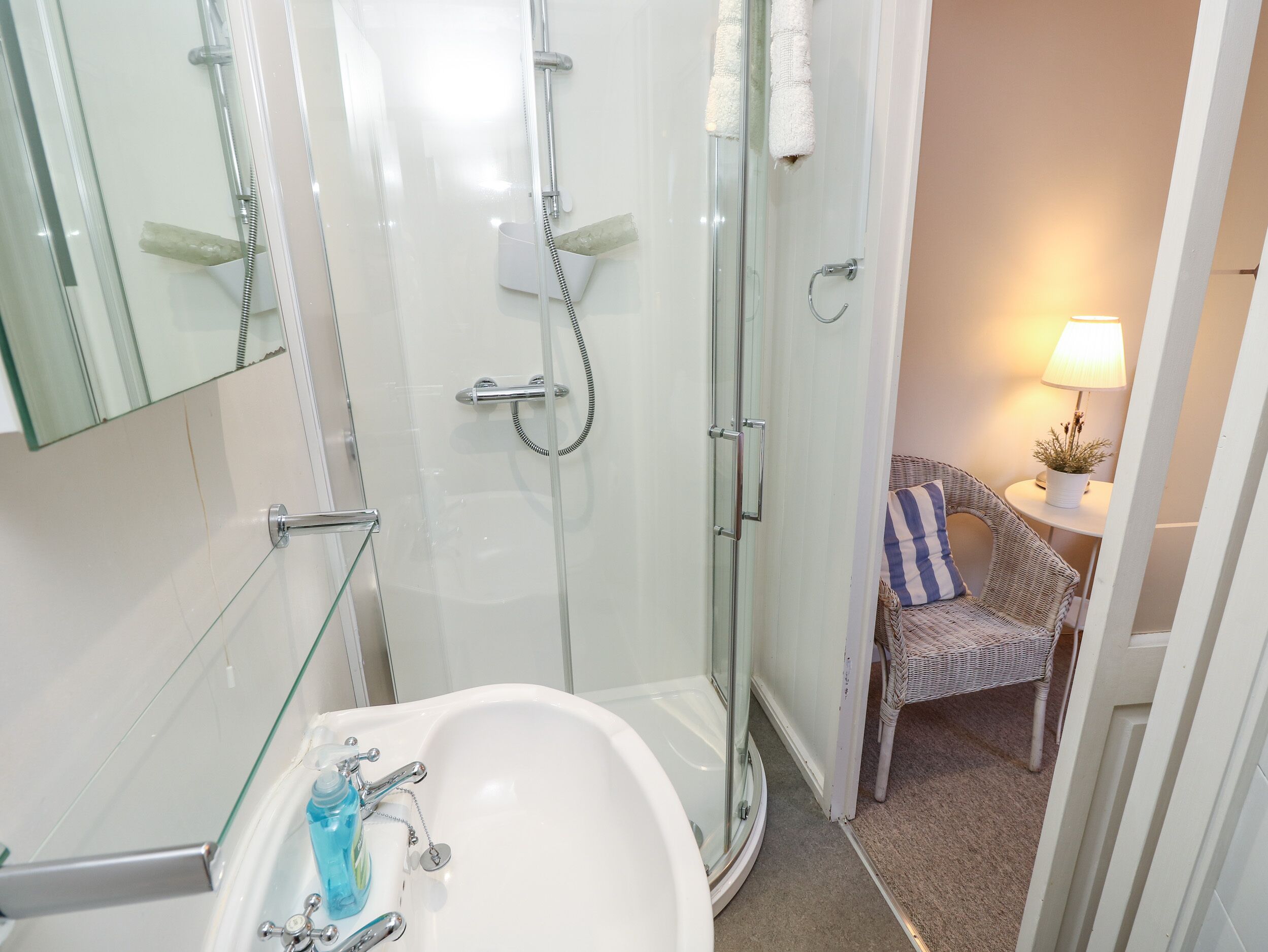 A bathroom with a glass-enclosed shower, a white sink with a glass shelf, and a wicker chair with a striped pillow.