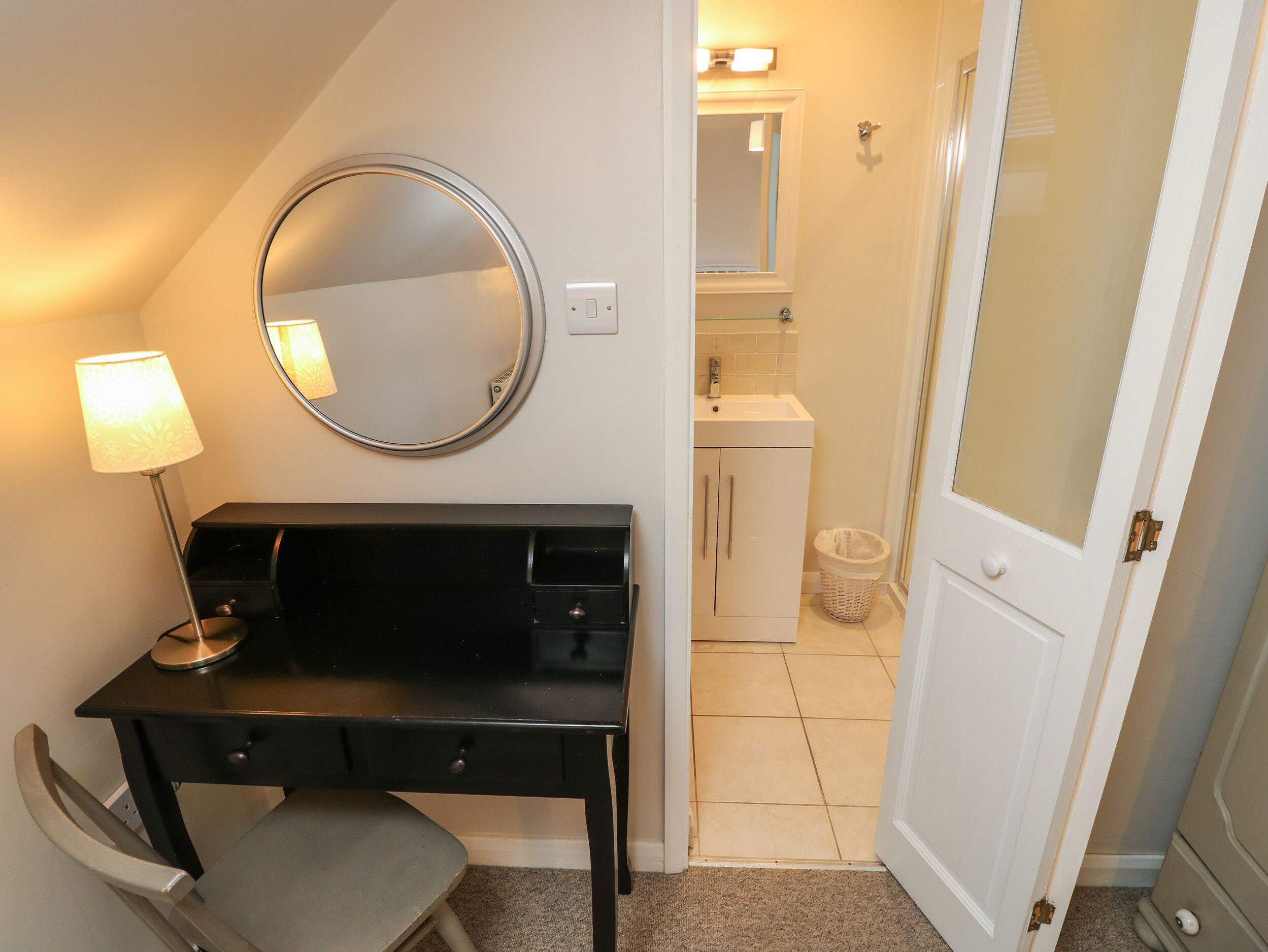 Habitación compacta con escritorio, silla y espejo. Se ve una puerta que conduce a un baño con lavamanos y un pequeño cubo de basura.