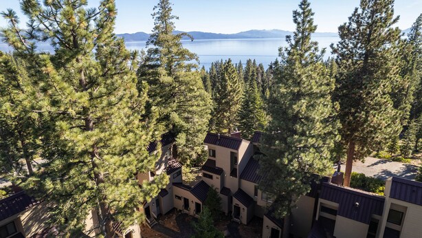 A residential area with multiple buildings, surrounded by tall pine trees, and a lake with mountains in the background. - Carnelian Woods # 126 by Hauserman Rental Group (Carnelian Bay)