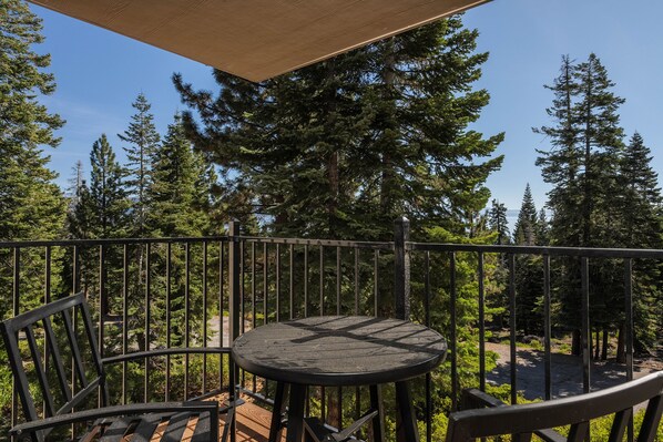 A balcony with a round table and chairs, surrounded by a metal railing, overlooking a forested area.