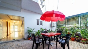 Um pátio externo com um guarda-sol vermelho, cadeiras pretas e vasos de plantas.