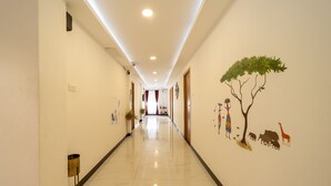 Corredor de hotel com piso de azulejos brilhantes, um mural na parede com uma árvore e animais, e uma porta no final.