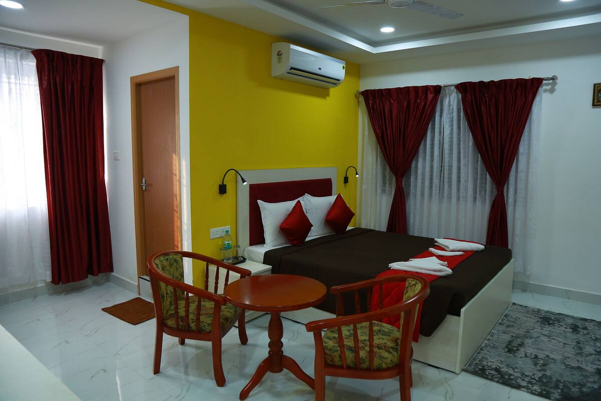 A hotel room with a bed, two chairs, a small table, an air conditioner, and a door.