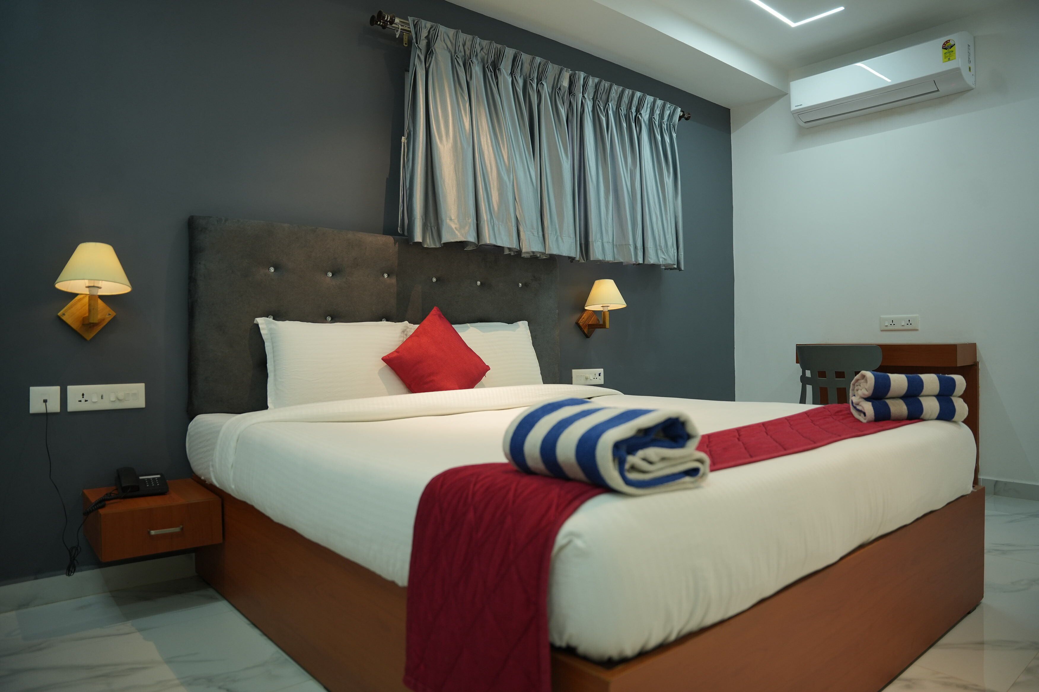 Habitación de hotel moderna con una cama grande, dos lámparas de noche, una mesita de noche, una colcha roja y una ventana con cortinas.