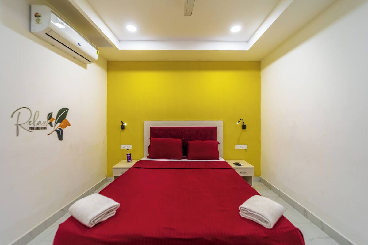 A hotel room with a red bedspread, two pillows, a white headboard, and a wall-mounted air conditioner.