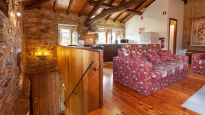 A rustic interior with stone walls, wooden beams, a wooden staircase, a floral sofa, and a kitchen area. - Loft Casinha - old barn in a rural area (Caminha)