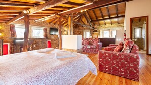A rustic room with exposed wooden beams, a stone wall, a bed with floral bedding, a red floral sofa, and a stone fireplace. - Loft Casinha - old barn in a rural area (Caminha)