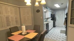 A dining area with a round table, four chairs, and a chandelier. A kitchen is visible in the background with a tiled backsplash and a refrigerator.
