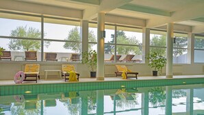 An indoor swimming pool with clear glass walls, wooden lounge chairs, and potted plants.