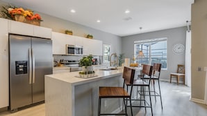 A modern kitchen with a stainless steel refrigerator, a microwave, and a sink. There are potted plants on the counter and a large window providing natural light.