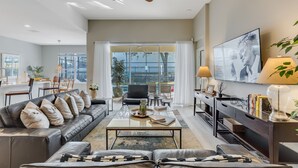 A modern living room with a leather sofa, a coffee table, and a TV. There is a dining area with a table and chairs, and a large window with curtains.