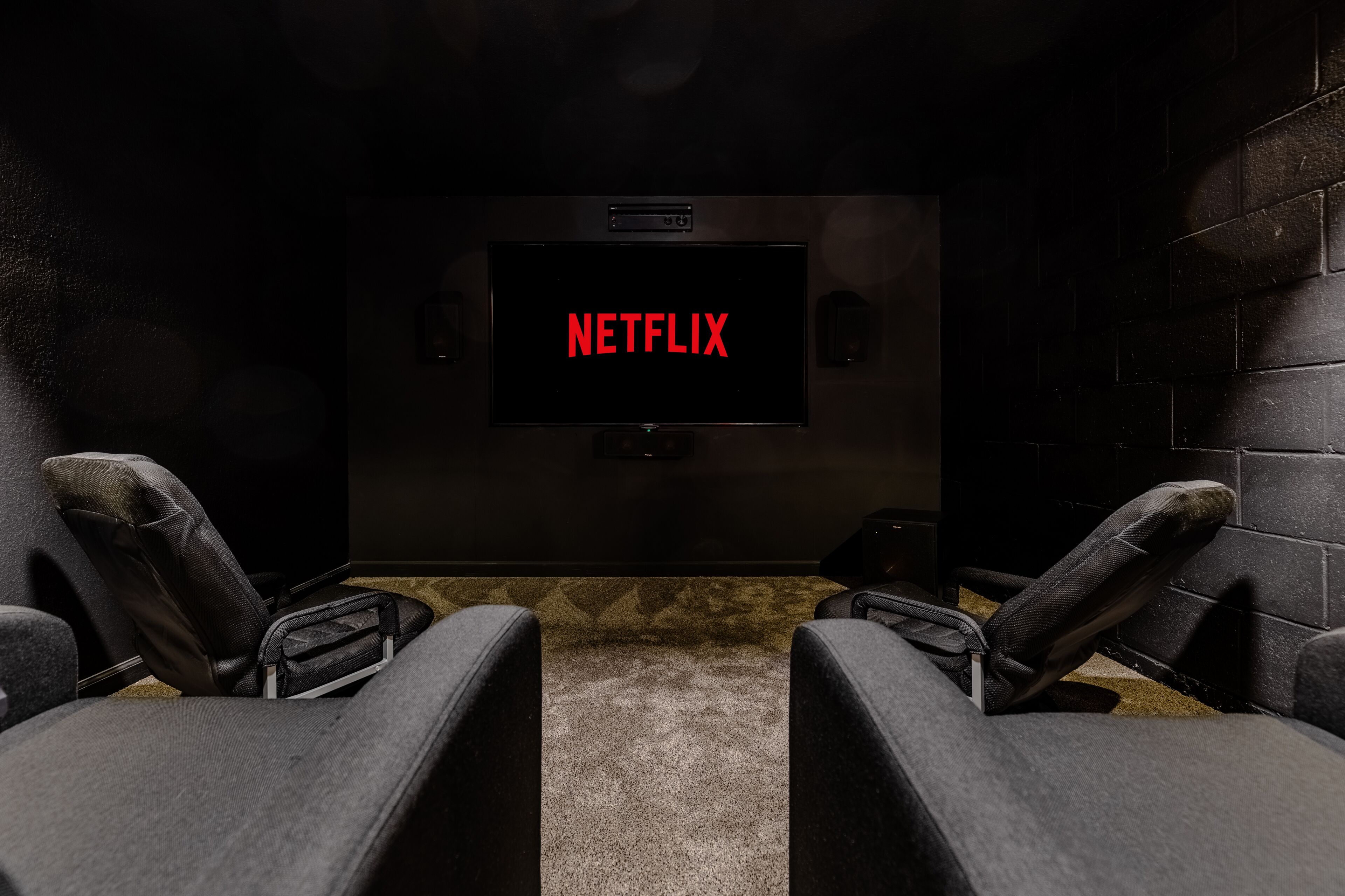 A dark room with a large screen displaying the Netflix logo, two reclining chairs, and a carpeted floor.