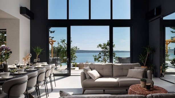 A modern interior with a large window overlooking a body of water, a sofa, dining table, and chairs.