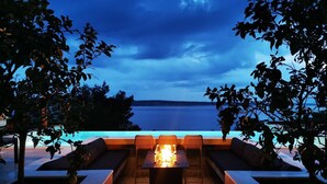 An outdoor seating area with a fire feature, surrounded by trees and overlooking a body of water.