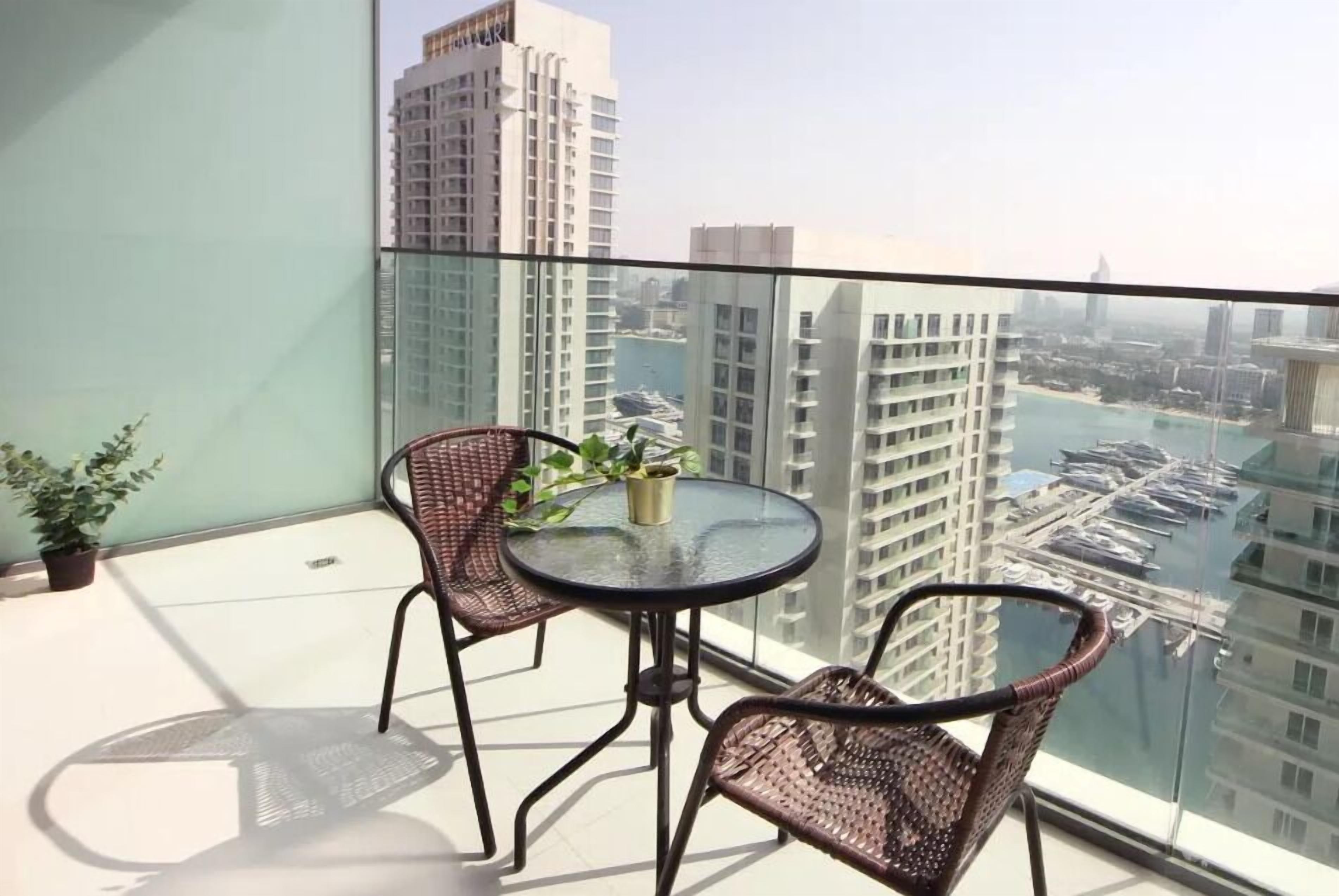 Een balkon met rieten stoelen en een glazen tafel, met uitzicht op een jachthaven en hoge gebouwen.