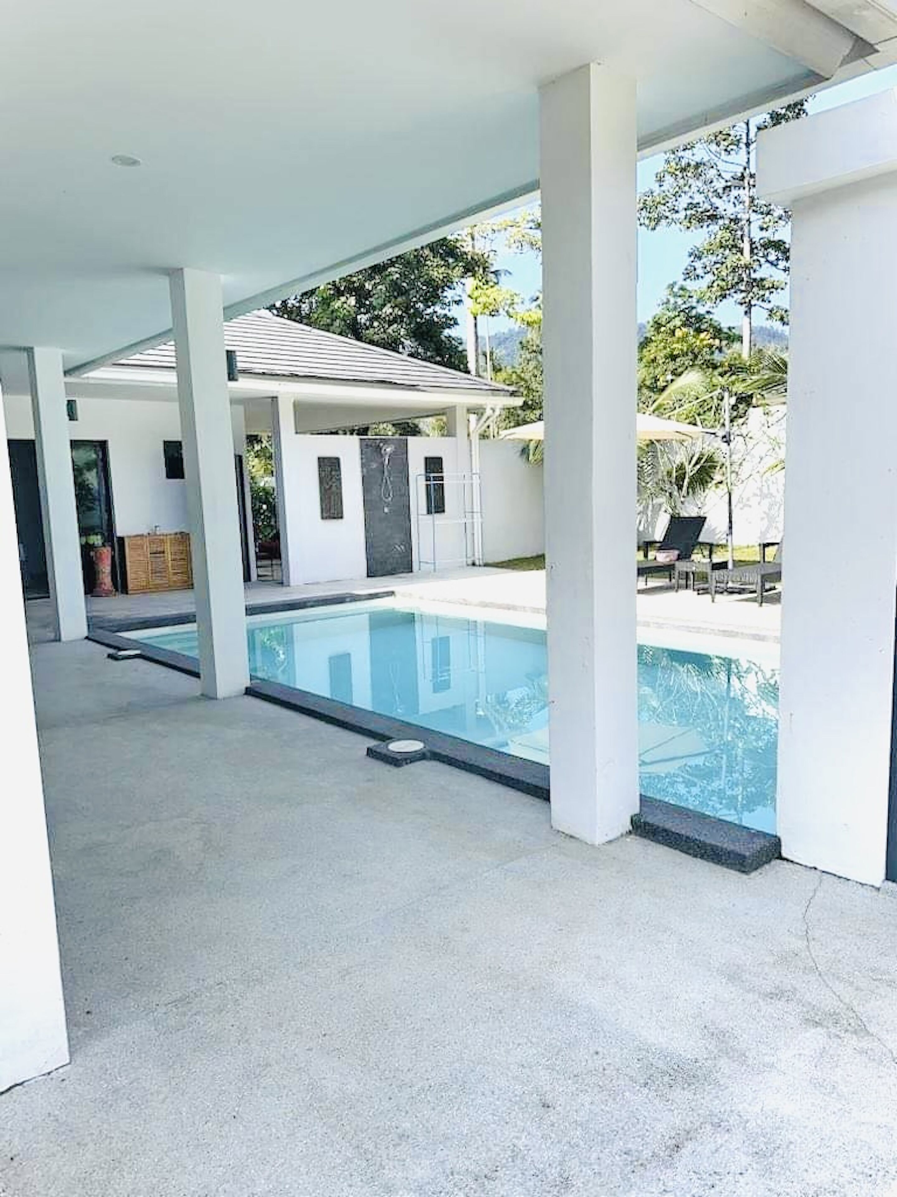 A covered swimming pool area with a clear view of the pool, a covered patio, and a building with a door and windows.