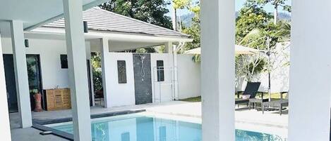 A covered swimming pool area with a clear view of the pool, a covered patio, and a building with a door and windows.