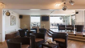 A lounge area with wicker furniture, a flat-screen TV, and a view of the city through large windows.