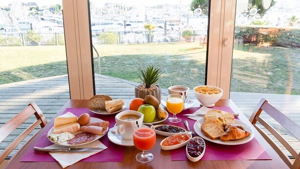 A breakfast table with various foods and drinks, set on a wooden deck with a view of a marina and buildings. - Le Vent du Large (Saint-Gilles-Croix-de-Vie)