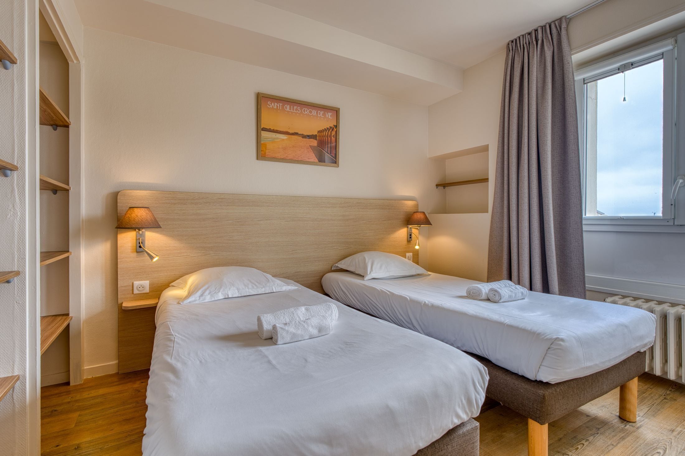 A hotel room with two beds, wooden headboards, bedside lamps, a poster on the wall, and a window with curtains.