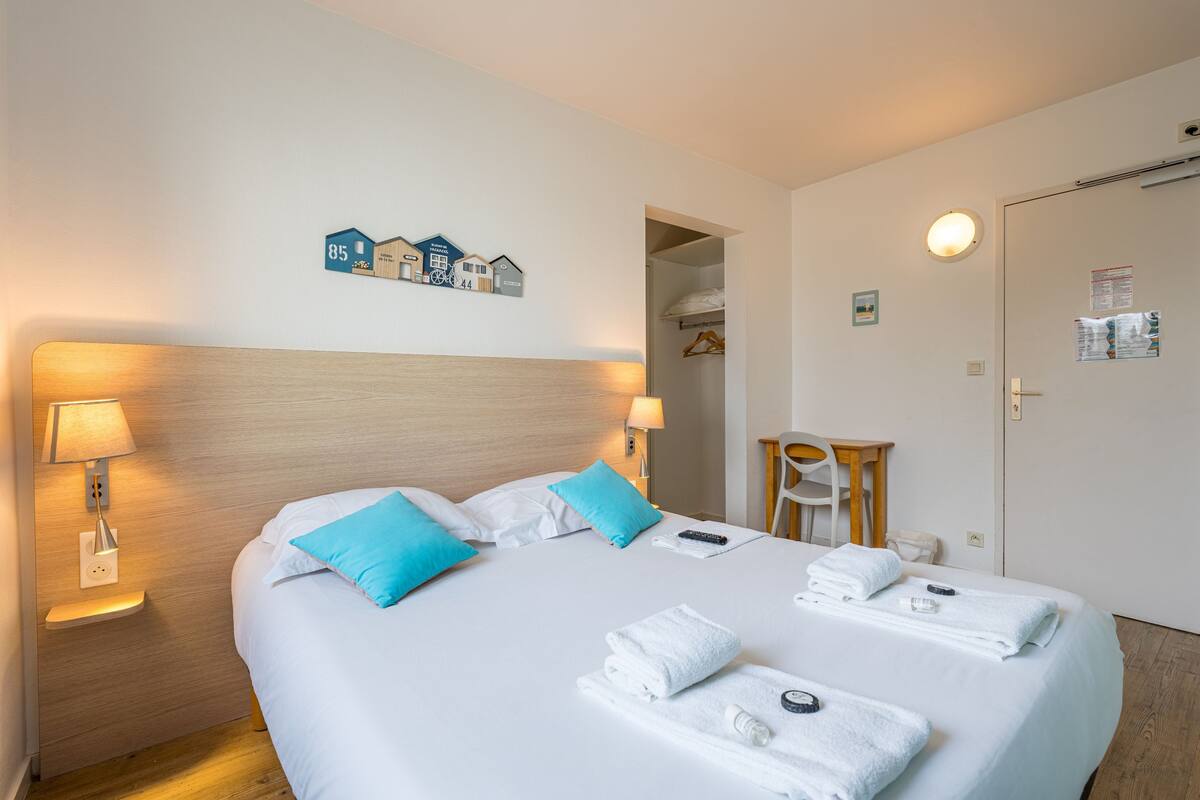 Una cama bien hecha con ropa de cama blanca y azul, dos lámparas de noche y un aplique de pared.