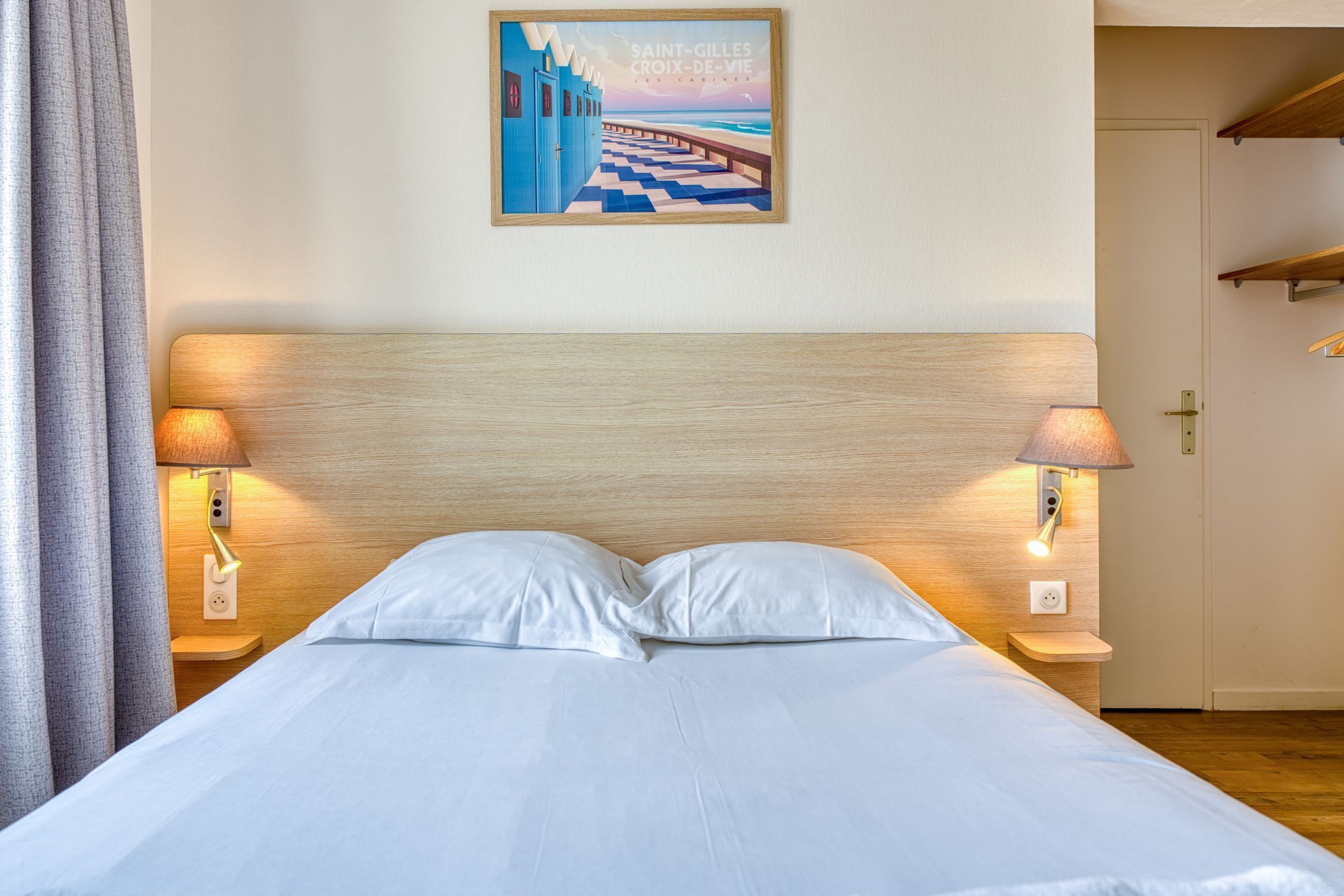 A hotel room with a bed, two bedside lamps, a wall-mounted picture, and a door.