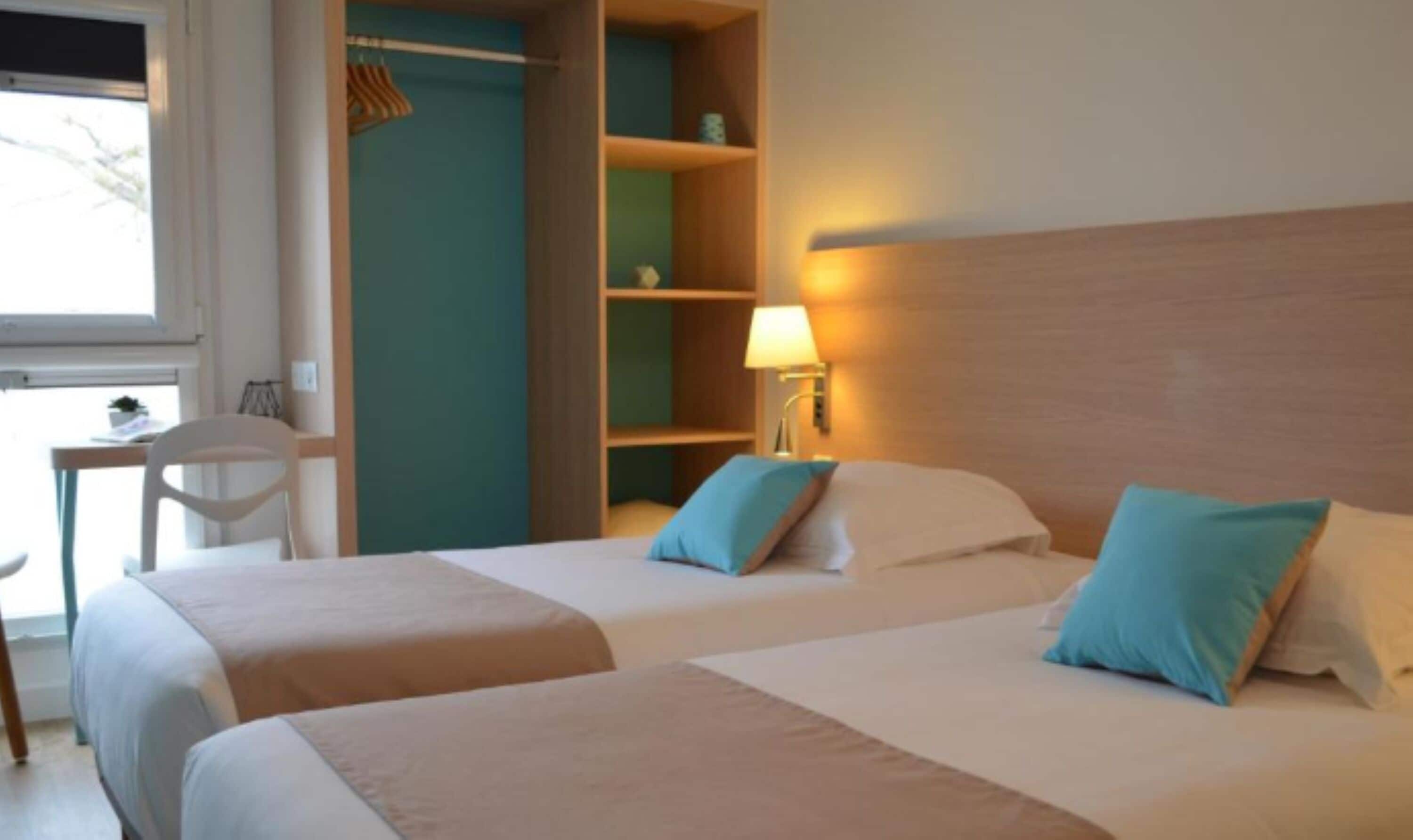 Habitación de hotel con dos camas, un escritorio y una silla. Se ve una ventana con vistas a árboles.