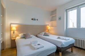 Habitación de hotel con dos camas, cada una con mesita de noche y lámpara, cabecero de madera, decoración de peces en la pared y ventana con vistas.