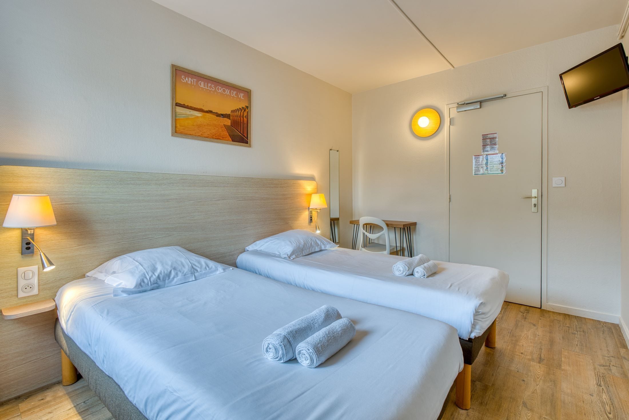 A hotel room with two beds, a TV, a poster, and a desk with a chair.