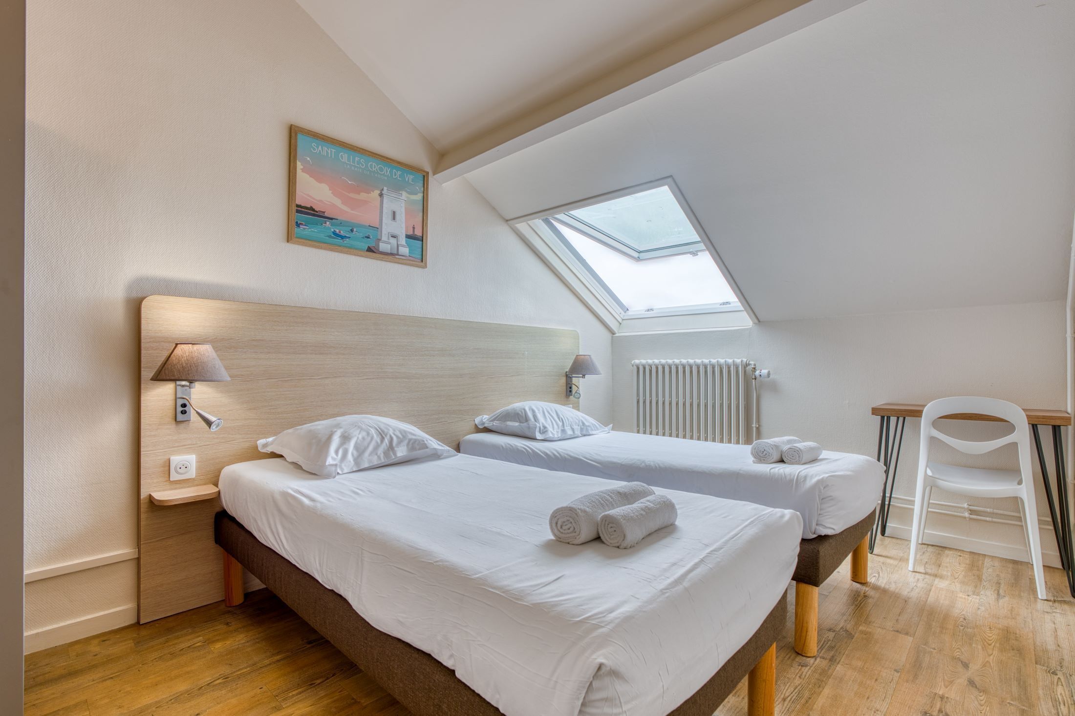 A bedroom with two beds, a skylight, a white chair, and a painting on the wall.