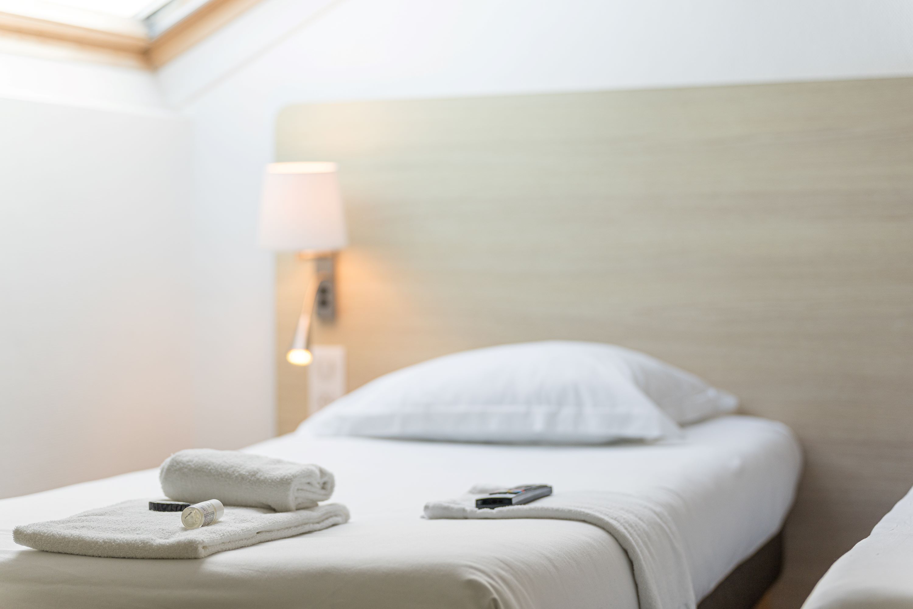 Una cama bien hecha con sábanas blancas, una toalla enrollada y un control remoto sobre la cama.