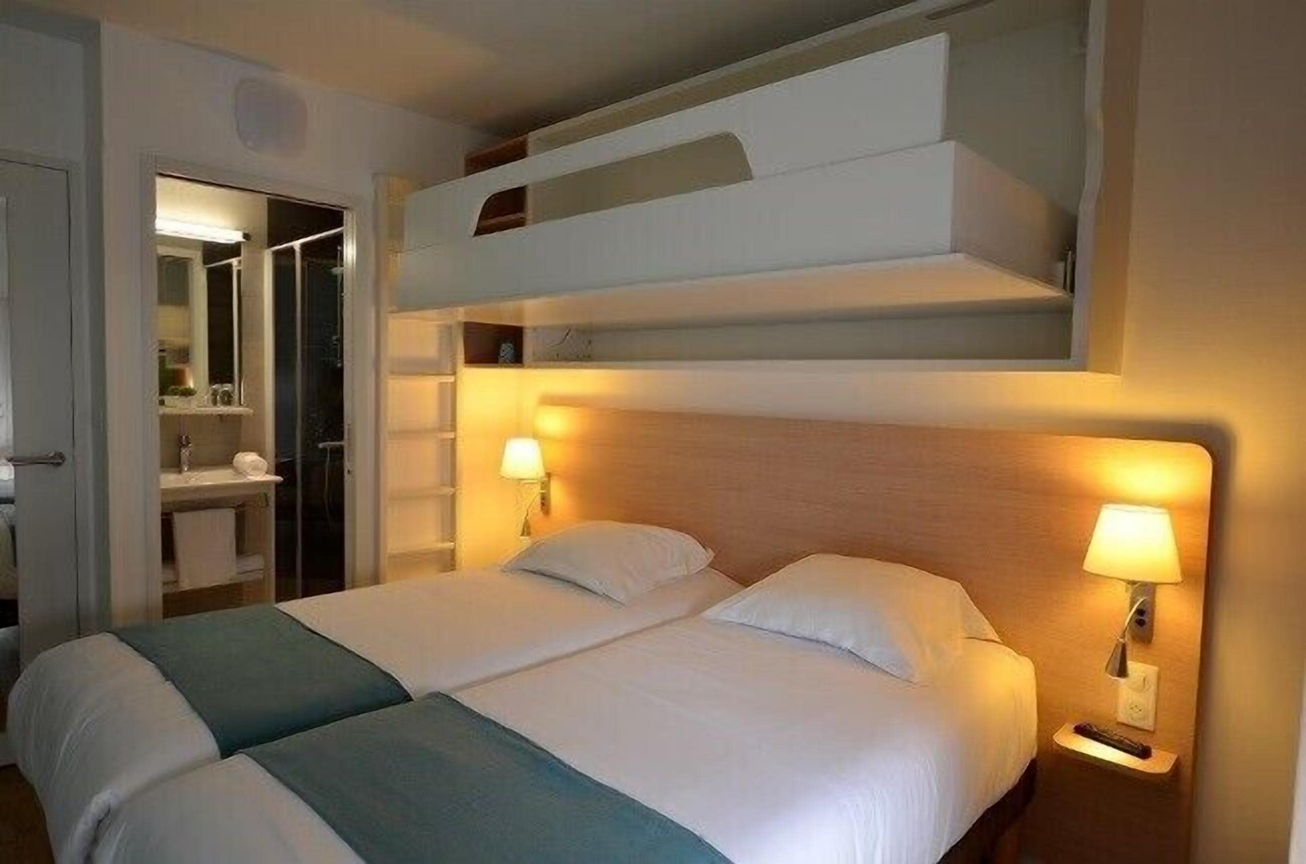 A hotel room with two beds, a bunk bed above, a bathroom with a mirror, and two bedside lamps.
