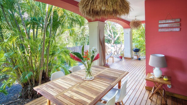 A wooden deck with a table and chairs, surrounded by tropical plants and a red wall. - Villas "les Saintes" Pour 4 à 5 Personnes à 50 m de la Plage au Calme (Saint-François)