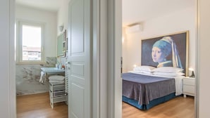 A bedroom with a bed, a nightstand, a painting, a window, and a bathroom with a sink and mirror.