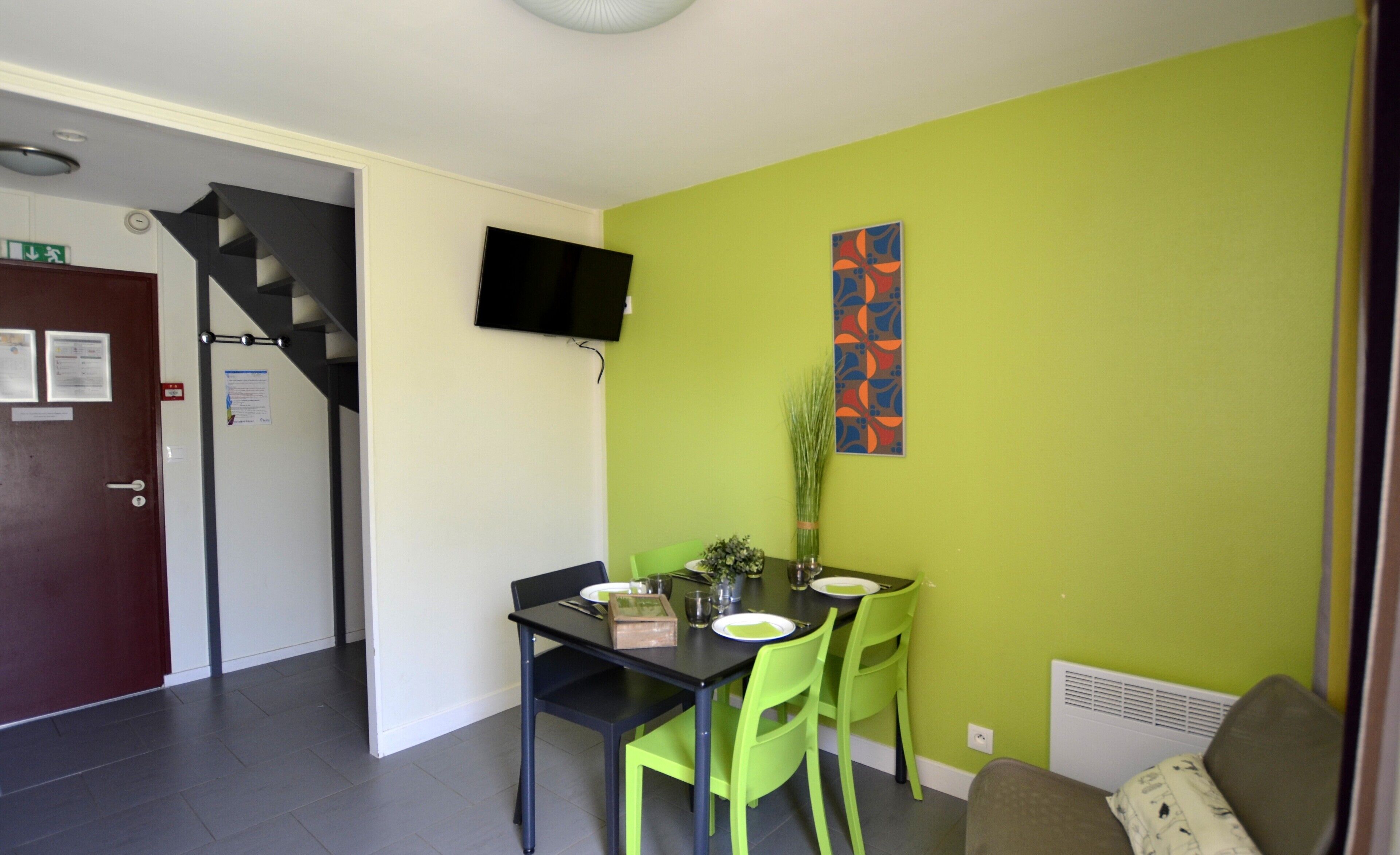 Un coin repas avec une table dressée pour deux personnes, deux chaises, un téléviseur fixé au mur et un escalier menant à une autre pièce.