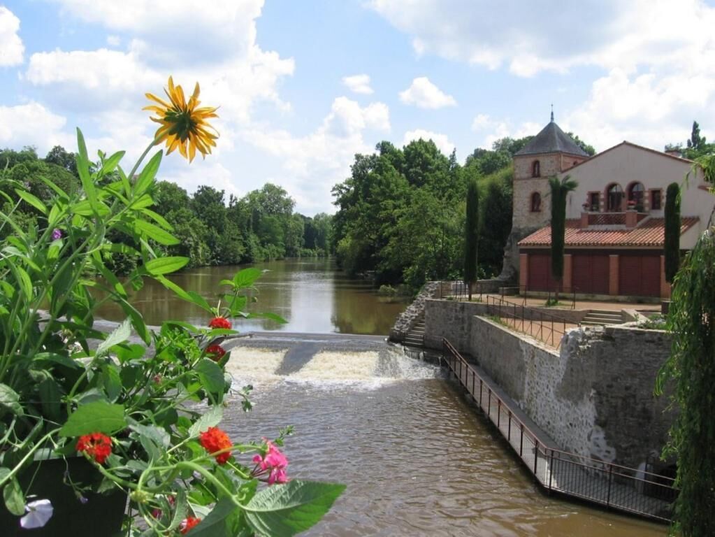 A river with a stone bridge, a building with a red roof, and greenery in the background.